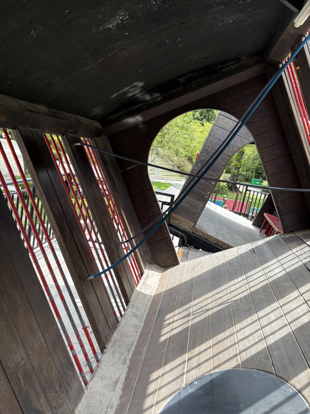 Inside a tilted carriage: cables stretched across the space at different angles, red bars along one side, a round opening looking out to the park, and a sloping wooden floor.