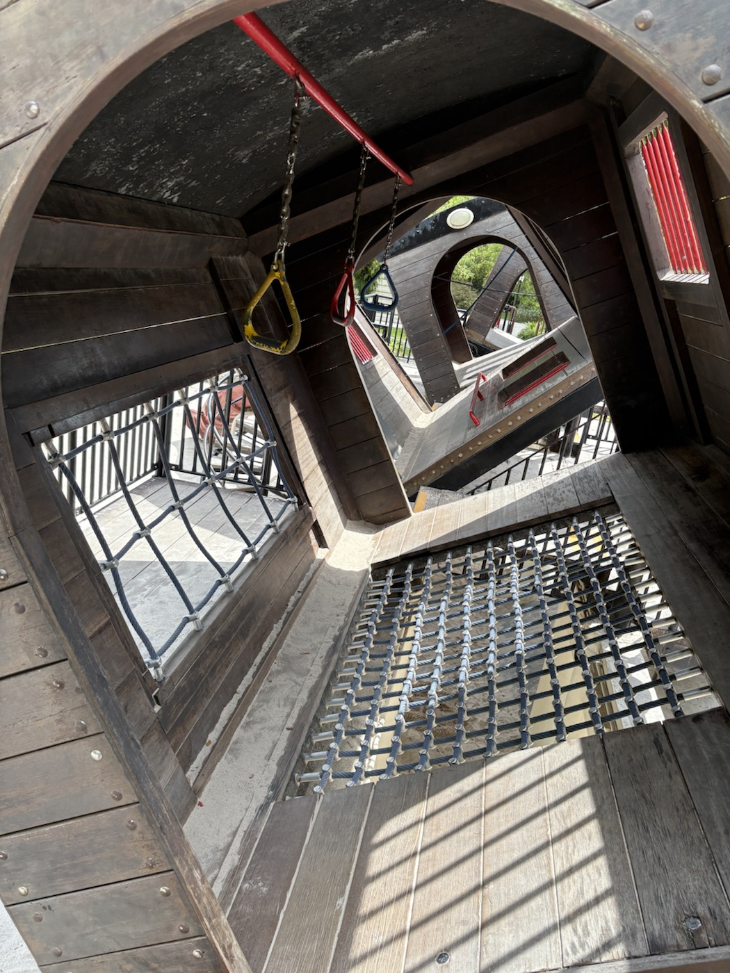 Inside a tilted wooden train carriage: three hanging rings on a red bar, a rope net window, and a rope net section in the floor, with sunlight coming through slats.