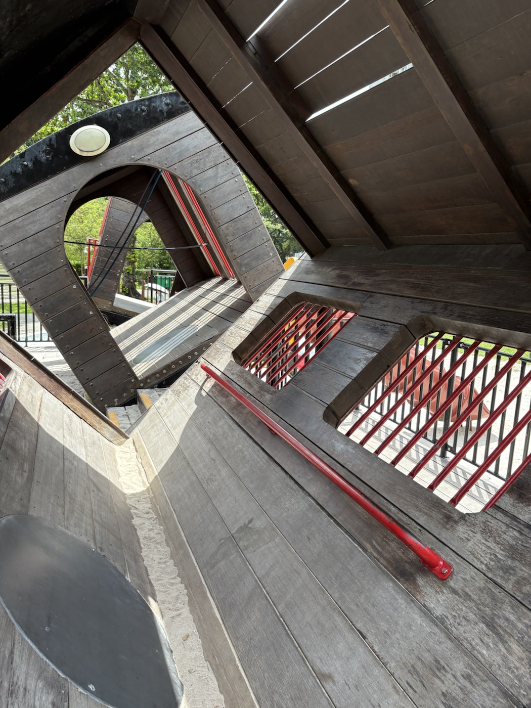 Inside another tilted carriage: wide sloping wooden boards, a red handrail running diagonally, red-barred windows and large round arched openings that frame the park.