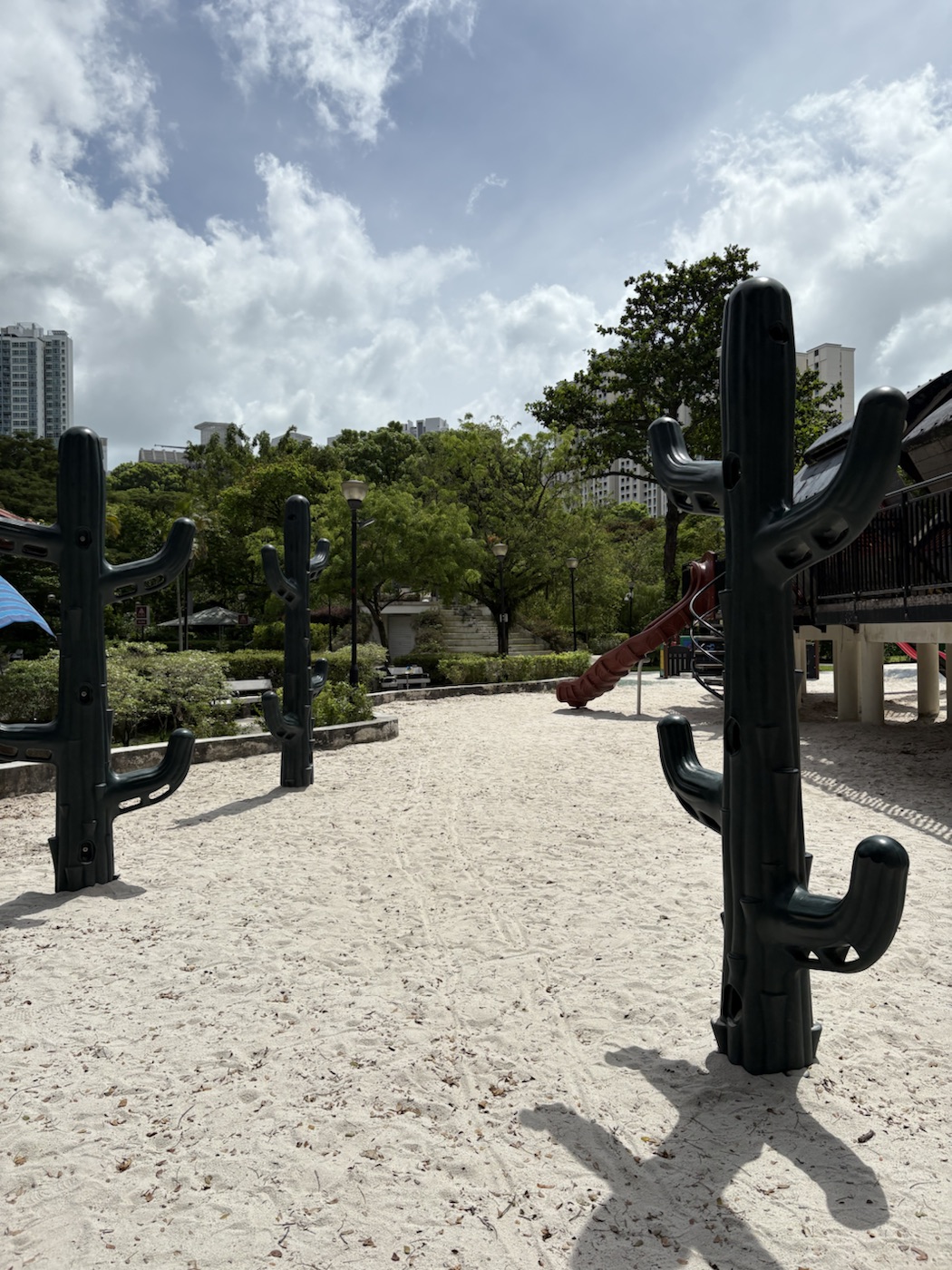 A wide stretch of pale sand with tall dark green cactus-shaped poles in the foreground and a train playground structure behind.