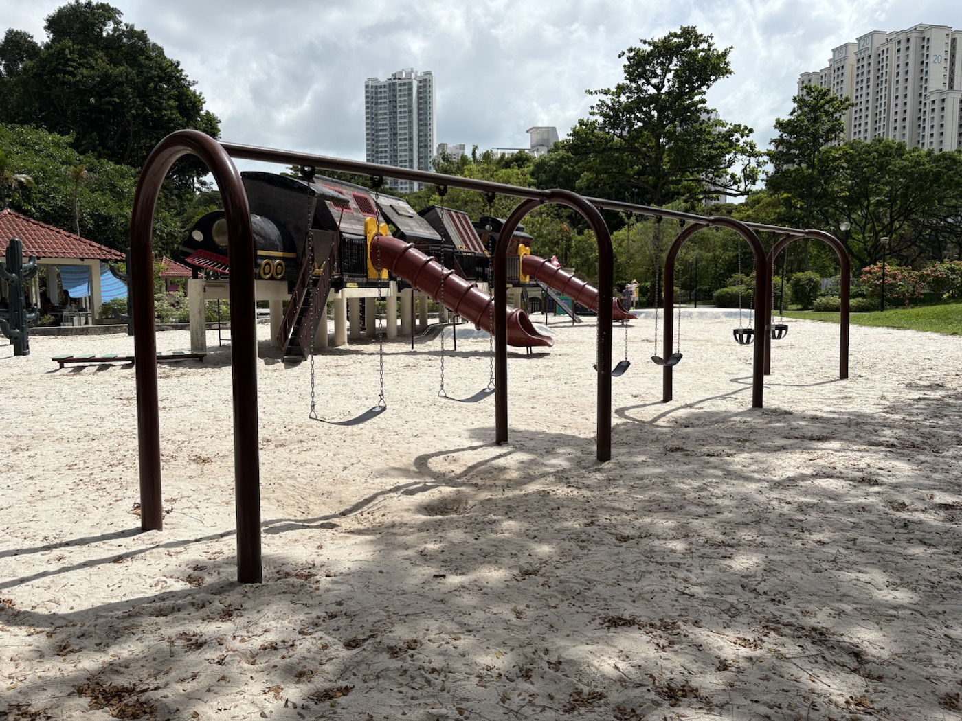 A long row of swings on a dark brown arched frame, with flat belt seats and two small bucket seats, set on pale sand in front of the train playground.