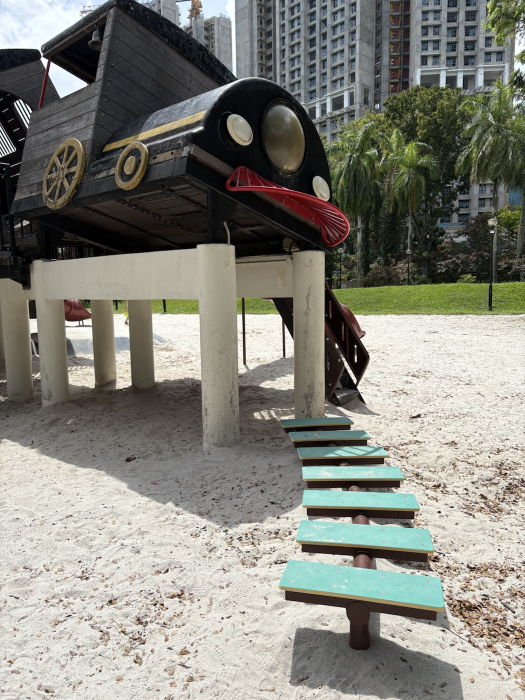 The front of the black train engine with round lamps and a red grille, raised on cream pillars, with a row of teal stepping planks on dark posts leading away across the sand.