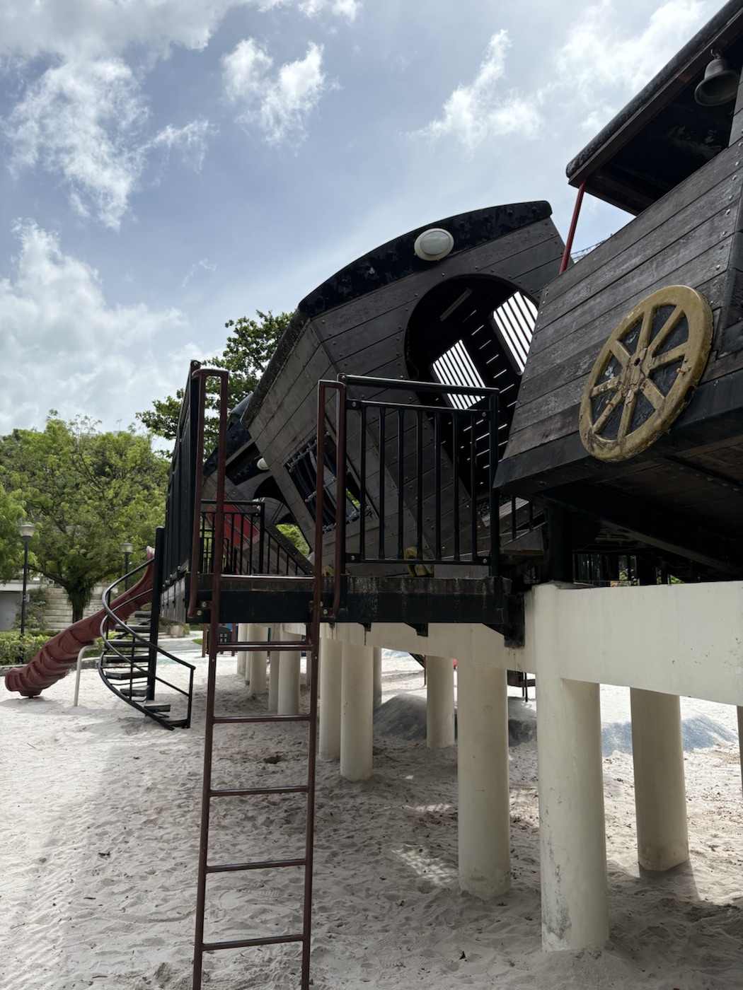 The side of the raised train carriages on cream pillars, with a tall brown metal ladder in the foreground, a spiral staircase and red curved slide on the left and a big yellow wooden wheel on the right.