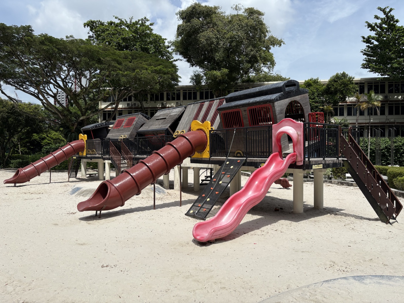 The whole train playground: a tilted black and red wooden train raised on pillars, with two long brown tube slides, a wavy pink slide, a black climbing ramp with coloured grips and a staircase on the right.