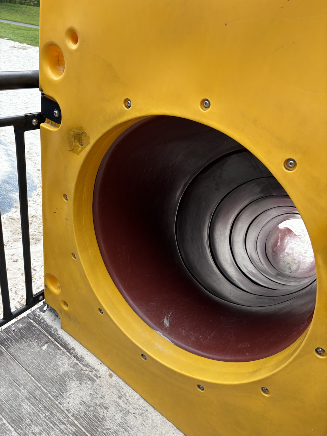 Looking into the mouth of a brown tube slide set in a yellow plastic panel; the tunnel curves away and a small circle of light shows at the far end.